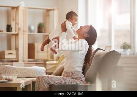 Vista laterale ritratto di felice afro-americano di madre holding simpatico baby boy mentre gioca in casa, spazio di copia Foto Stock