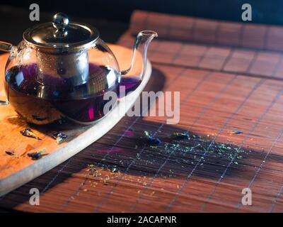 Tè tailandese blu in una teiera di vetro. Tè appena preparato, bevanda calda deliziosa e sana. Bevanda calda su Mat, vita morta scura. Foto Stock