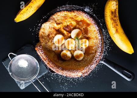 Concetto di cibo in casa baby olandese o tedesco banana Topping Caramel crêpe in una padella di ferro su sfondo nero Foto Stock