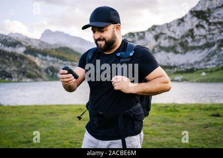 Uomo caucasico con cappuccio e barba utilizza il GPS in un paesaggio di montagna con un lago Foto Stock