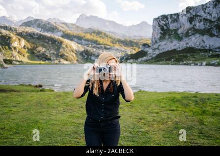 Una bella donna caucasica con un abbigliamento informale consente di scattare fotografie con una fotocamera in un paesaggio di montagna con il lago Foto Stock