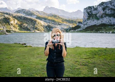 Una bella donna caucasica con un abbigliamento informale consente di scattare fotografie con una fotocamera in un paesaggio di montagna con il lago Foto Stock