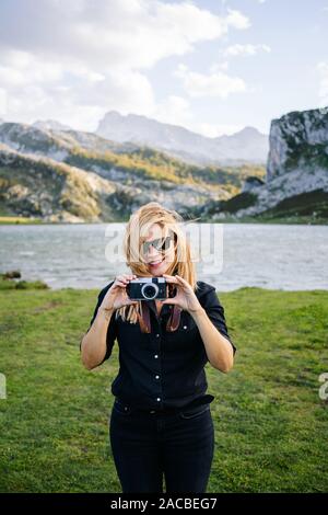 Una bella donna caucasica con un abbigliamento informale consente di scattare fotografie con una fotocamera in un paesaggio di montagna con il lago Foto Stock