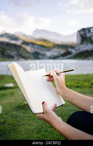 Una bella caucasian donna bionda scrive e disegna su un blocco note in un paesaggio di montagna con il lago Foto Stock