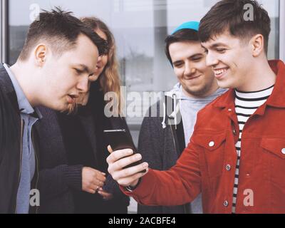 Giovane uomo che mostra qualcosa di divertente sul suo smartphone a un gruppo di adolescenti amici, concentrarsi sulla mano che tiene il telefono cellulare e il filtro opaco Foto Stock