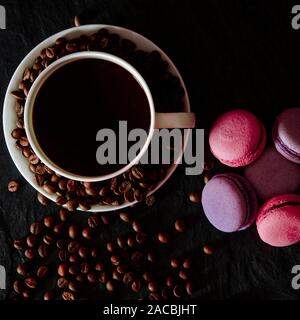 Una tazza di bianco con il nero fragranti caffè sorge su uno sfondo nero con caffè zurnes accanto al francese colorati cookies amaretti Foto Stock