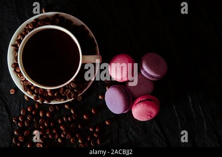 Una tazza di bianco con il nero fragranti caffè sorge su uno sfondo nero con caffè zurnes accanto al francese colorati cookies amaretti Foto Stock