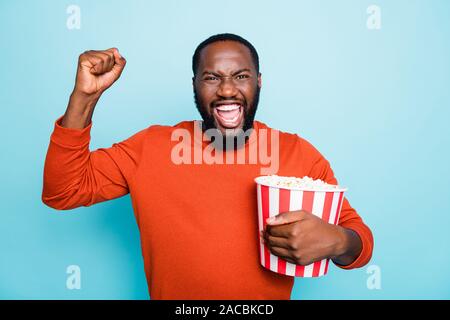 Foto di allegro contentissimo estasiato uomo pazzo urlando in felicità tenendo i pop corn con benna emotivo espressione facciale in arancione maglione Foto Stock