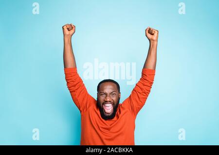 Foto di incredibile crazy pelle scura guy alzando i pugni gridare a gran voce il supporto di squadra di calcio preferita casual wear orange pullover isolato di colore blu Foto Stock