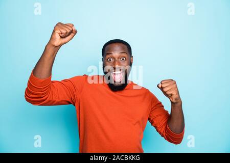 Foto di incredibile crazy pelle scura guy celebrando vincente della lotteria alzando i pugni urla trionfante usura arancione informale pullover isolato di colore blu Foto Stock