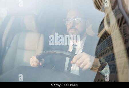 Giovane imprenditore barbuto in occhiali tenendo la ruota e la guida della vettura in città Foto Stock