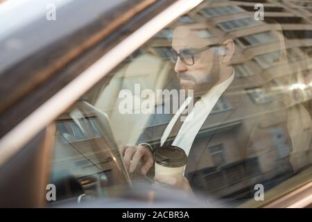 Giovane imprenditore in occhiali lavorando su laptop e di bere il caffè mentre è seduto nella sua auto Foto Stock