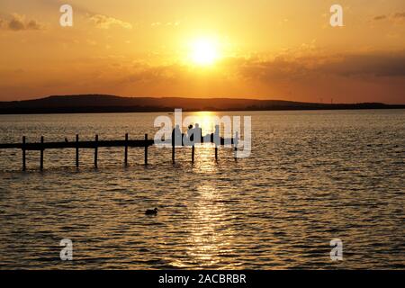 Gruppo di amici rilassante sul molo a guardare il tramonto sul lago Steinhuder Meer in Germania, irriconoscibile la gente in silhouette Foto Stock