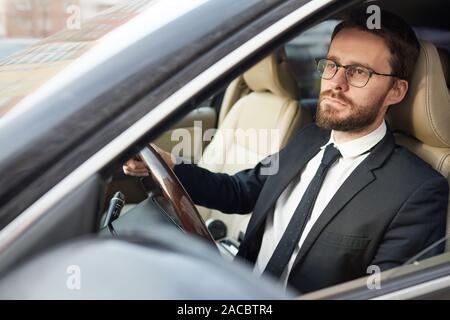 Grave giovane imprenditore barbuto in occhiali seduto al volante e guidare la vettura ha concentrandosi sulla strada Foto Stock