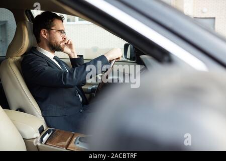 Grave imprenditore barbuto seduto in auto di lusso e parlando al telefono cellulare durante la guida della macchina Foto Stock