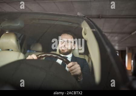 Giovane uomo gravi in occhiali tenendo la ruota e la seduta nella sua auto nel parcheggio Foto Stock