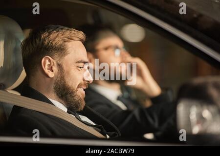 Vista laterale del giovane passeggero barbuto sorridere mentre è seduto in macchina con autista alla guida della vettura Foto Stock