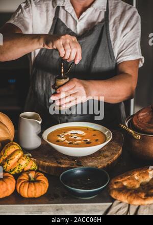 La donna in grigio grembiule mettendo il pepe alla crema di zucca zuppa Foto Stock