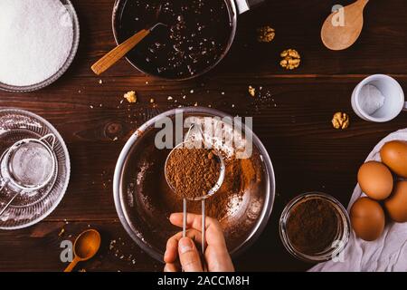 Femmina vagliatura a mano il cacao in polvere nel recipiente di metallo accanto agli ingredienti per fatti in casa torta brownie: cioccolato fuso, noci, zucchero, farina e uova o Foto Stock