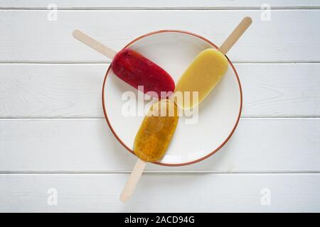 Vista aerea di tre diversi tipi di frullato di frutta o popsicles pop di ghiaccio su una piastra bianca su sfondo di legno Foto Stock