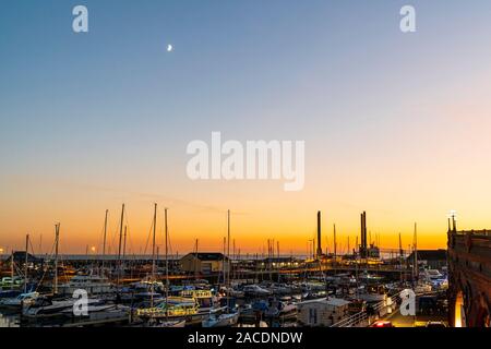 Il cielo giallo diventa blu dopo il tramonto al porto di Ramsgate, nel Kent, in Inghilterra. Primo piano, piccole barche e yacht, molti illuminati con luci di Natale, nel porticciolo nel porto interno, e attraverso i molti alberi di yacht, una banda di cielo giallo e arancione dopo il tramonto. Foto Stock