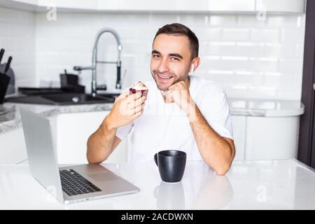 Giovane uomo chiamando sul telefono cellulare tramite airpods mangiare torte mentre si lavora sul portatile a casa al mattino Foto Stock