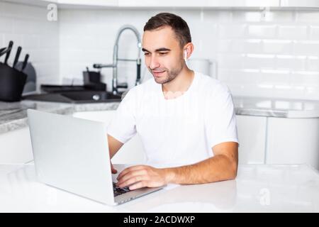 Uomo informale chiamando sul telefono cellulare tramite airpods mentre si lavora sul computer portatile a casa al mattino Foto Stock