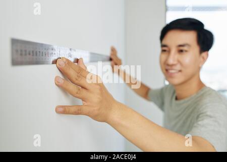 Sorridente giovane uomo utilizzando metallo ruller quando si misurano le pareti del suo appartamento Foto Stock
