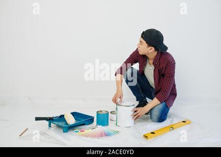 Pensieroso giovane uomo nel cappuccio guardando blank muro bianco nel suo nuovo appartamento durante l'apertura di latta di vernice Foto Stock