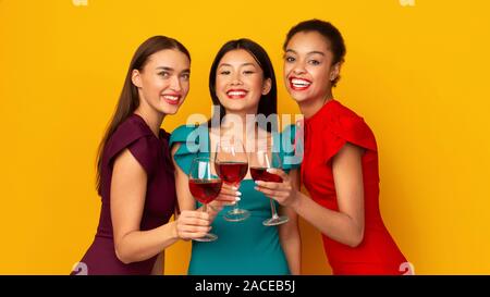 Tre ragazze con vino rosso bicchieri tintinnanti, sfondo giallo, Panorama Foto Stock