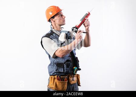 Un builder in un casco con silicone in una pistola di montaggio. Tiene in mano. Su uno sfondo luminoso. Foto Stock