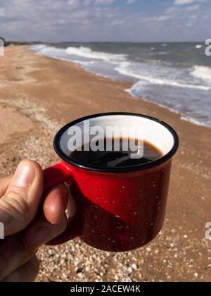 In viaggio con il tè. Close up man mano azienda red mug con scuri puerh tea con le onde del mare e spiaggia vuota sulla copia di sfondo spazio vicino fino Foto Stock