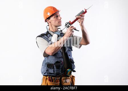 Un builder in un casco con silicone in una pistola di montaggio. Tiene in mano. Su uno sfondo luminoso. Foto Stock