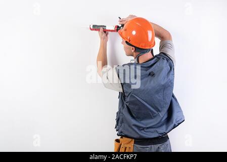 Un builder in un casco con silicone in una pistola di montaggio, mette sulla parete. Su uno sfondo luminoso. Foto Stock