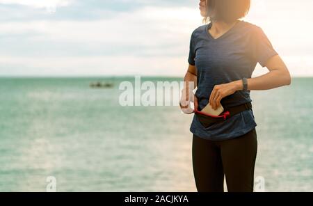 Giovane donna asiatica tenere lo smartphone nel marsupio prima di eseguire cardio esercizio al mattino alla spiaggia del mare. Allenamento all'aperto. Runner e smart usura di banda Foto Stock