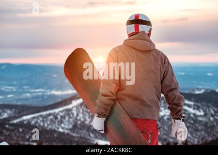 Snowboarder si erge con lo snowboard in mani contro il tramonto e le montagne Foto Stock