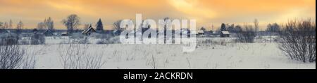 Panorama invernale con un bellissimo tramonto sopra il villaggio russo e la coperta di neve campo sotto un cielo arancione Foto Stock