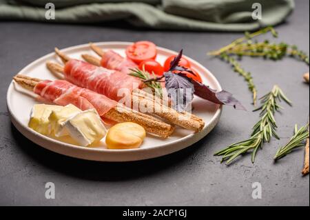 La cucina italiana tradizionale è pane grissini con prosciutto e formaggio e pomodori con erbe su una piastra su uno sfondo scuro. Close up Foto Stock