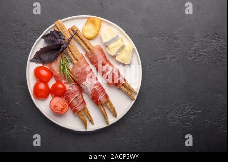 La cucina italiana tradizionale è pane grissini con prosciutto e formaggio e pomodori con erbe su una piastra su uno sfondo scuro. Un sano stile. Spazio di copia Foto Stock