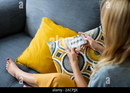 Chiudere fino sulla donna di mani tenendo lo smartphone con il logo Amazon. Giovane donna usando il telefono con Amazon logo sullo schermo a casa accogliente sul divano in camera Foto Stock