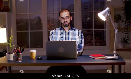 Imprenditore barbuto che indossa una camicia e prendere un sorso di caffè mentre si lavora da casa di notte ore. Foto Stock