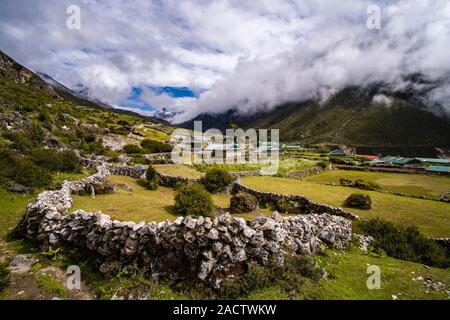 I campi del piccolo villaggio circondato da mura di pietra, paesaggio montuoso coperto di nuvole di monsone a distanza Foto Stock