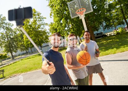 Sport e tempo libero giochi e amicizia maschile concetto - gruppo di uomini felici o amici di scattare una foto da smartphone su selfie stick su outdoor basketball pl Foto Stock