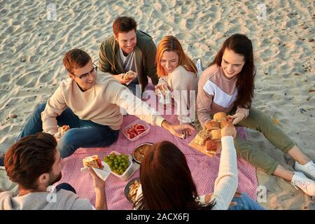Amicizia, di svago e di fast food concept - gruppo di felice amici mangiando panini o hamburger a Picnic sulla spiaggia in estate Foto Stock