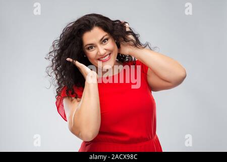 Persone, bellezza e ritratto concetto - donna felice in abito rosso di toccare i suoi capelli su sfondo grigio Foto Stock
