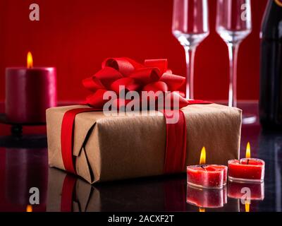 Primo piano di un regalo di San Valentino e il cuore ardente di candele sagomato con bicchieri di champagne in background in base a una tabella di scuro e uno sfondo rosso Foto Stock