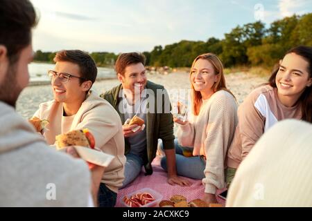 Amicizia, di svago e di fast food concept - gruppo di felice amici mangiando panini o hamburger a Picnic sulla spiaggia in estate Foto Stock