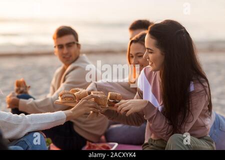 Amicizia, di svago e di fast food concept - gruppo di felice amici mangiando panini o hamburger a Picnic sulla spiaggia in estate Foto Stock