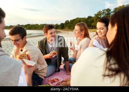 Amicizia, di svago e di fast food concept - gruppo di felice amici mangiando panini o hamburger a Picnic sulla spiaggia in estate Foto Stock