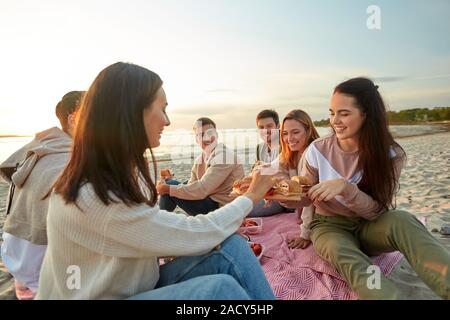 Amicizia, di svago e di fast food concept - gruppo di felice amici mangiando panini o hamburger a Picnic sulla spiaggia in estate Foto Stock